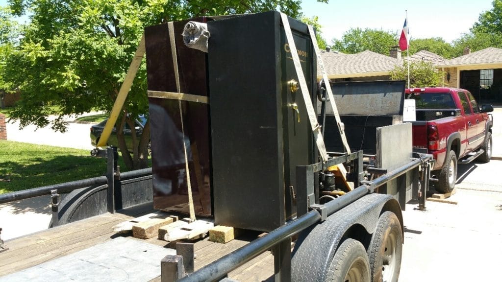 Large black safe strapped on a trailer hitched to a red pickup truck in a suburban driveway.