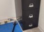 Black four-drawer filing cabinet with metal handles and a lock, placed in a corner of a room on a light floor.